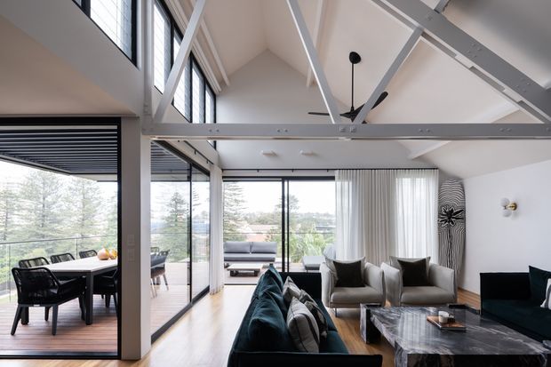 A bright, open-plan living room with a dining area visible to the left, expansive floor-to-ceiling glazing, and views onto a timber deck and landscaped surroundings. The room features a vaulted ceiling with exposed white structural beams, a large ceiling fan, light timber flooring, neutral upholstered armchairs, dark sofas, and a substantial dark stone coffee table. Vertical sheer curtains and dark window frames create a contemporary indoor-outdoor connection.