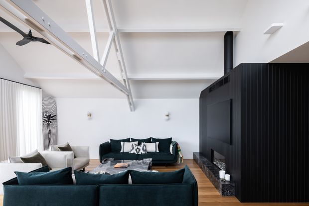 Bright contemporary living room with a large deep teal sectional sofa facing a matching dark sofa and a black built-in fireplace or media wall. The room has white walls and ceiling, exposed white-painted structural beams, tall sheer-curtained glazing, warm timber flooring, and a striking full-height black vertical-slat feature wall. A black flue rises to the ceiling, while restrained monochrome furnishings and artwork create a refined, high-contrast interior.