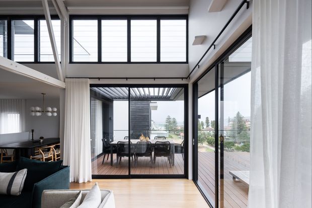 Bright contemporary open-plan living and dining area with expansive black-framed glazing, sheer curtains, light timber flooring, and a view onto a covered timber deck and outdoor dining area. A partial kitchen or dining zone is visible to the left, while the room is defined by its indoor-outdoor connection and high clerestory-style windows.