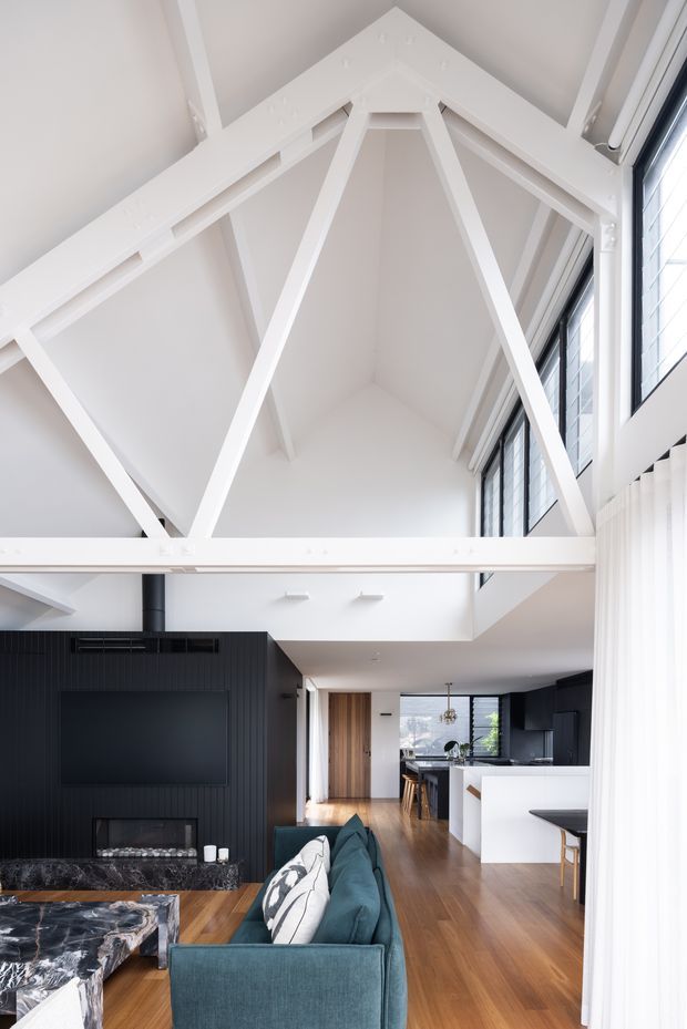 Bright double-height open-plan living space with a dramatic white exposed timber truss roof structure and tall black-framed windows. The room includes a dark vertically panelled fireplace and media wall, teal sectional sofa, marble coffee table, timber flooring, and a partially visible contemporary kitchen and dining area beyond.