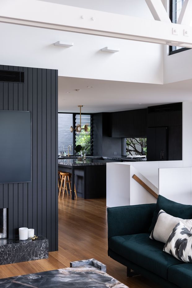 A contemporary open-plan living area with a dark vertically panelled feature wall and integrated fireplace or media unit, opening into a compact black kitchen with a marble-look island benchtop and timber flooring. A deep teal sofa occupies the foreground beside a dramatic dark stone coffee table. White walls, a stair enclosure with timber handrail, pendant lighting, and high-level glazing create a bright, high-contrast interior with a partially double-height feel.