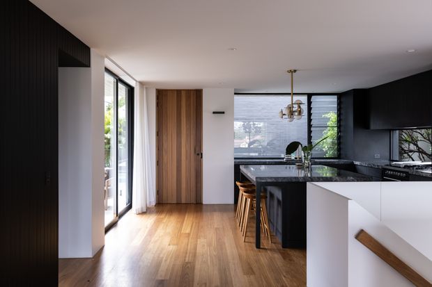 A contemporary open-plan kitchen viewed from the adjacent entry or living area. The room features warm timber flooring and a vertical timber door, a black island and dark cabinetry, white walls and white island panels, large glazed doors, and a sculptural pendant light. A staircase with a timber handrail is visible in the foreground.