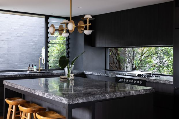 A dark, sophisticated contemporary kitchen with a large central island, integrated cooktop, extensive black cabinetry, and dark polished stone surfaces. Warm timber bar stools sit beneath the island overhang, while brass-and-glass pendant lighting adds contrast. Horizontal windows bring in greenery and daylight, and a pale brick wall is visible through the glazing.