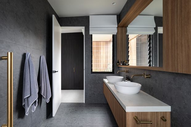 A refined contemporary bathroom with dark charcoal wall and floor finishes, a long timber vanity with a pale stone benchtop, two white vessel basins, brass wall-mounted tapware, and a large mirrored cabinet. A glass shower enclosure is visible at left, while a doorway opens to a dark-finished adjoining space. The room has strong linear detailing, warm timber accents, and soft natural light through windows with Roman blinds.