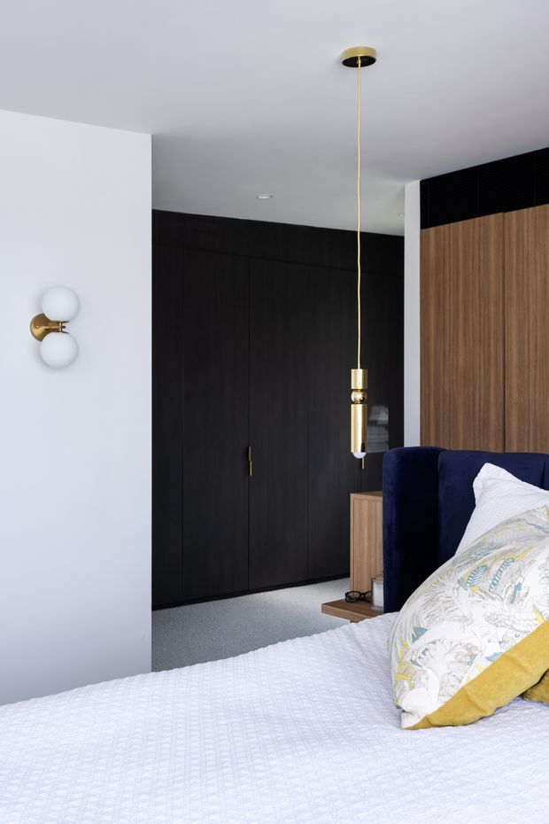 A refined contemporary bedroom with a large bed in the foreground, upholstered in white bedding with patterned cushions and a mustard accent pillow. The room features a deep navy upholstered headboard, dark timber built-in wardrobes, a lighter timber bedside cabinet, and a white wall with a sculptural brass-and-glass wall sconce. A slender brass pendant light hangs beside the bed, creating a sophisticated contrast with the dark joinery and soft neutral finishes.