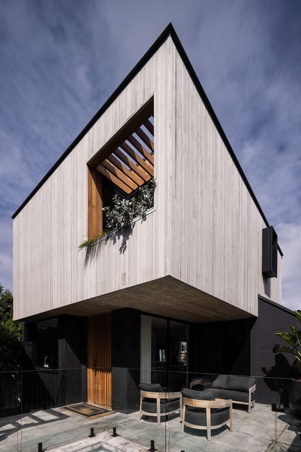 A contemporary two-storey house exterior with a sharply pitched gable roof and vertically textured light cladding. The upper level features a dramatic recessed opening with exposed timber rafters and greenery, while the dark lower level opens onto a paved terrace furnished with outdoor lounge chairs and a sofa behind a glass balustrade. Strong black trim and dark masonry contrast with the pale facade.