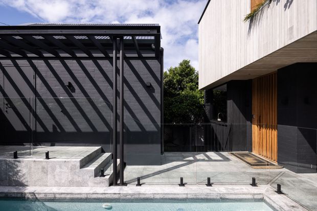 Contemporary residential exterior courtyard beside a swimming pool, featuring dark rendered walls, a slim black metal pergola, pale concrete paving and steps, a light vertical-cladding upper volume, and a warm timber entry screen. Strong linear shadows and clean geometric forms create a polished modern outdoor setting.