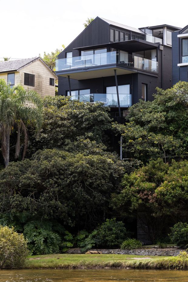 Exterior view of a contemporary multi-level dark-clad residence set within dense landscaped greenery beside a body of water. The house features a steep gable roof, broad projecting balconies with clear glass balustrades, extensive glazing, slim dark structural supports, and an adjoining flat-roofed volume. A smaller weatherboard house is visible to the side.