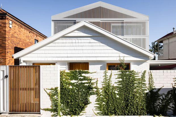 Front exterior view of a compact white masonry house with a prominent pitched gable roof and a modern, vertically screened upper structure behind it. The facade includes white painted brickwork, horizontal shingle-like cladding on the gable, timber-look vertical slatted gates, timber-framed openings, and a planted boundary wall. Neighbouring brick and weatherboard buildings are visible on either side.