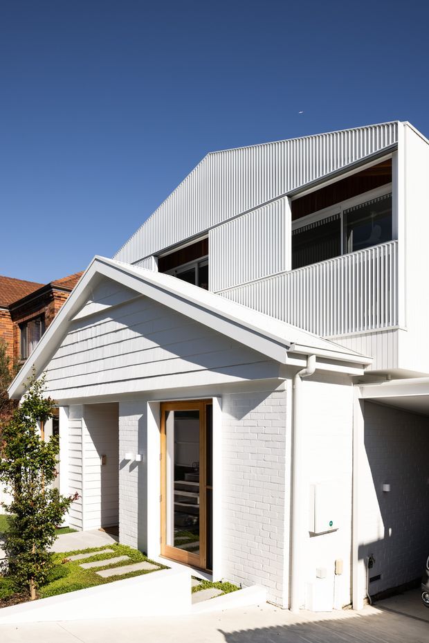 Bright exterior view of a contemporary multi-level urban residence. The white-painted brick lower level has a steep gabled entry volume with horizontal cladding, a timber-framed glazed front door, and a narrow planted path. Above, the addition features white vertical ribbed cladding and matching vertical screening around large dark-framed windows and a balcony. A covered driveway or garage opening is visible at the right, with a neighbouring brick roof partially visible behind.