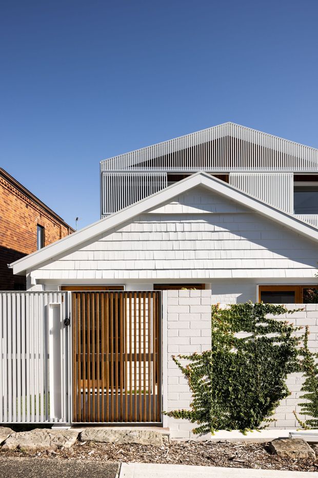 Front exterior view of a compact residential house with a white-painted gabled facade, textured shingle-like cladding, white masonry walls, a timber-slat entry gate, and a modern upper-level addition screened by closely spaced vertical slats. A neighbouring red-brick building is visible at the left edge under a clear blue sky.