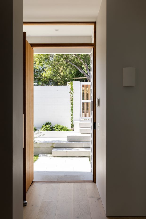 View from a narrow interior passage through an open timber-framed doorway toward a bright landscaped courtyard. The space has pale timber flooring, white walls, a simple wall light, and a contemporary pivot or hinged door with warm wood framing. Outside, stepped pale paving, raised planters, a white masonry boundary wall, and a timber-slatted gate are visible. A glazed transom above the doorway brings additional daylight into the passage.