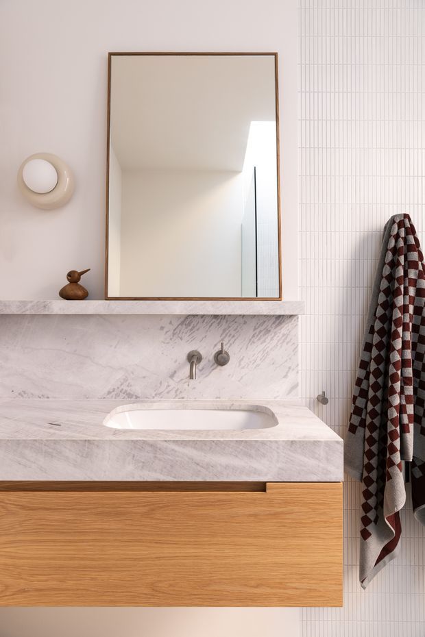 A refined bathroom vanity composition featuring a wall-mounted timber vanity with a white marble or stone benchtop and integrated basin. The wall behind is clad in narrow vertical white tiles. A large rectangular mirror with a slim timber frame is mounted above the vanity, alongside a sculptural round wall light. Brushed metal wall-mounted tapware, a small decorative object, and a patterned burgundy, white, and charcoal towel complete the scene.