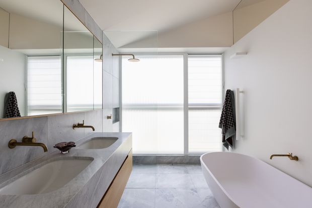 Bright contemporary bathroom featuring a double vanity with a pale stone benchtop, wall-to-wall mirror, large-format grey floor and wall tiles, a freestanding white bathtub, and a glass-enclosed shower. Brass wall-mounted tapware and warm neutral finishes add refined contrast. Roller blinds diffuse daylight through the large windows.