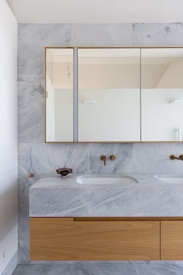 A contemporary bathroom vanity with twin integrated white basins set into a pale grey-veined stone benchtop and matching large-format stone wall and floor tiles. The wall-mounted timber vanity has flat-front drawers, while a large mirrored cabinet with a slim warm timber frame spans the wall above. Brushed brass or bronze wall-mounted tapware provides a subtle accent.