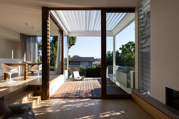Bright contemporary open-plan living and dining area opening onto a timber deck through large dark-framed glazed doors. A white louvred pergola provides cover over the outdoor area, with built-in outdoor cabinetry or a barbecue visible to the right. The interior includes pale timber flooring, a neutral palette, a dining table with modern chairs, a low built-in fireplace surround, and strong indoor–outdoor connection with views toward neighbouring homes and trees.