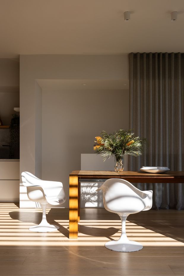 A refined dining area with a rectangular timber dining table, two sculptural white pedestal chairs, and a glass vase of foliage and orange flowers. Warm sunlight casts strong linear shadows across the timber floor and furniture. The room has a neutral plastered interior, full-height dark grey curtains, recessed ceiling lighting, and a large opening toward an adjacent space.