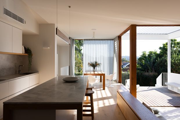 Bright open-plan kitchen and dining area opening onto a covered timber deck. The space features minimalist white cabinetry, a large dark stone island, timber flooring and joinery, full-height glazing with vertical blinds, and warm timber-framed openings to the outdoor area. Lush greenery and neighbouring rooftops are visible beyond the deck.