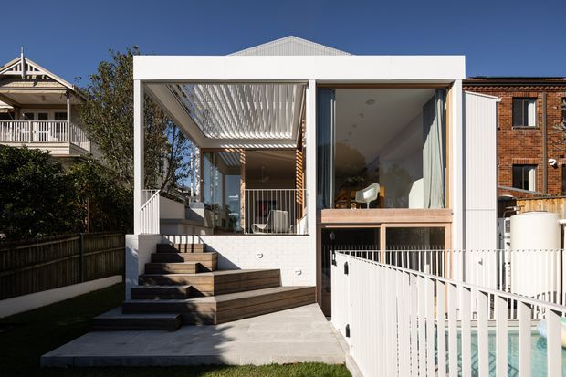 Exterior view of a contemporary two-storey residential extension in an urban neighbourhood. The white rectangular facade has large glazed openings, a covered upper terrace with a slatted pergola, timber-clad stair and deck elements, and white metal balustrades. A swimming pool is visible in the foreground, with neighbouring traditional and brick homes at either side.