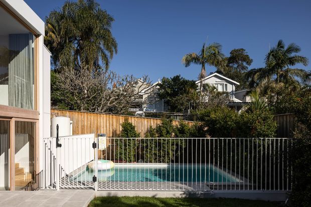Sunny suburban residential exterior with a fenced swimming pool in the foreground, timber boundary fencing, tropical and subtropical planting, and neighbouring pitched-roof homes behind. A contemporary house edge with large glazed openings is visible at left. The scene has a clear blue sky and a bright, relaxed outdoor setting.