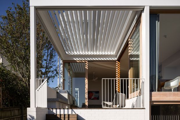 Exterior view of a contemporary residential terrace or balcony beneath a white-framed louvred pergola. The covered outdoor area has white metal balustrades, a rendered or painted brick base, timber decking or steps, and extensive glazing connecting to the interior. Strong linear shadows from the roof louvers define the space, with a glimpse of modern interior furnishings through the windows.