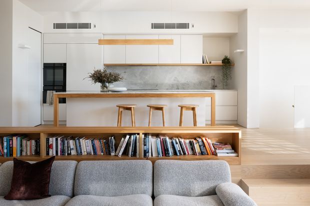 Bright open-plan kitchen and living area with a minimalist built-in kitchen, pale cabinetry, a grey stone-look splashback, timber breakfast bar and stools, and an integrated low bookshelf separating the kitchen from a grey sofa. Light timber flooring and white walls create an airy, refined interior.