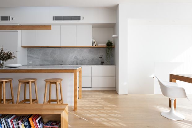 Bright open-plan kitchen and dining area with minimalist white cabinetry, a grey stone-look splashback and benchtop, light timber flooring, and a timber island with three stools. A partially visible dining table and white pedestal chairs occupy the adjoining space. Integrated linear ventilation grilles, simple shelving, and restrained styling create a clean contemporary interior.