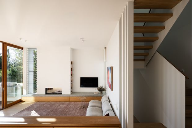 Bright contemporary living room with warm timber built-in joinery and a long low media console, a neutral sofa, wall-mounted television and expansive glazing opening toward greenery. An open-riser timber staircase with white vertical balustrade slats occupies the right side, creating a strong architectural feature. The room has a calm, refined palette of white, warm wood and muted grey-brown tones.