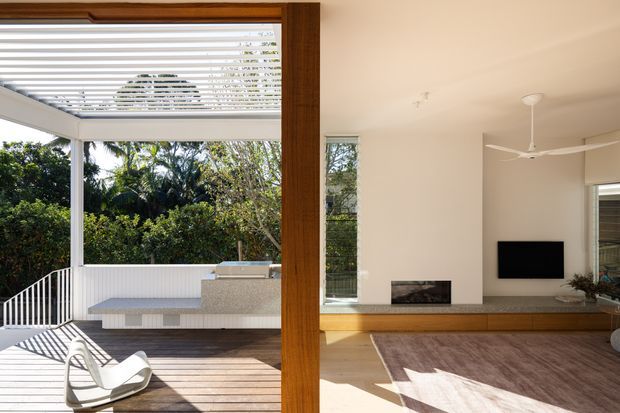 Bright contemporary living room opening onto a timber deck and covered outdoor entertaining area. The interior features pale walls, warm timber flooring and joinery, a linear fireplace, wall-mounted television, ceiling fan, and large glazing. Outside, a built-in tiled bench or outdoor kitchen sits beneath a louvred roof, surrounded by dense greenery.