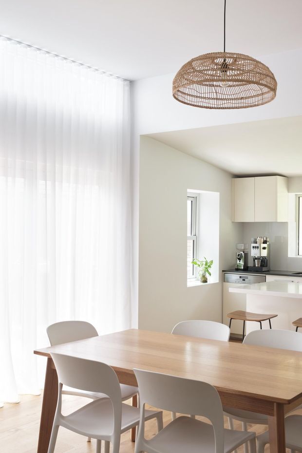 Bright open-plan dining area adjoining a compact kitchen. A light timber dining table with modern white chairs sits beside full-height sheer curtains, while a woven pendant light hangs above. The kitchen features pale cabinetry, a light benchtop, integrated appliances and a small breakfast counter with stools. The overall interior is airy, warm and minimalist.
