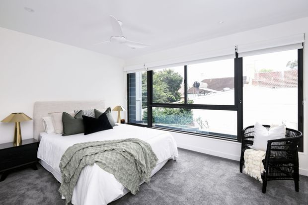 Bright contemporary bedroom with a neatly made bed, layered white and grey bedding, dark accent cushions, bedside tables and brass lamps. Large black-framed windows provide abundant natural light, while grey carpet and white walls create a calm, neutral interior. A woven black armchair with a light throw sits beside the glazing.