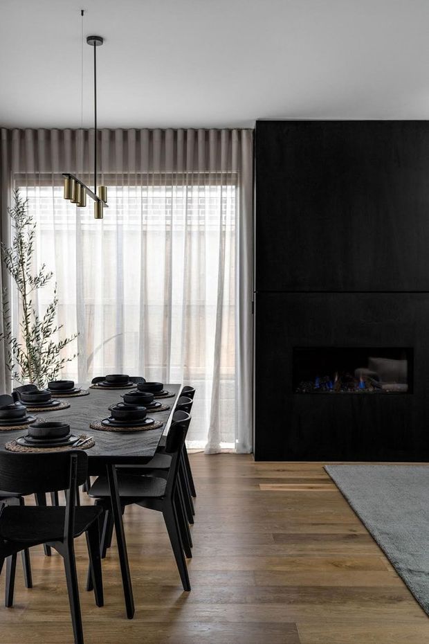 A refined open-plan dining and living area with a dark sculptural fireplace wall, timber flooring, floor-to-ceiling sheer curtains, and a black dining table set with dark tableware. A linear pendant light hangs above the dining table, while a neutral rug and part of the living area are visible to the right. The space has a restrained, contemporary monochrome palette with warm timber accents.