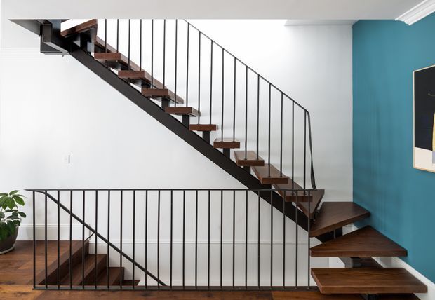 A bright modern stairway with floating dark timber treads supported by a black steel stringer and slender vertical balustrades. The stair turns at a landing beside a bold teal-blue feature wall, with white walls and ceiling and timber flooring or stair surfaces. A small potted plant and framed wall artwork are visible at the edges.