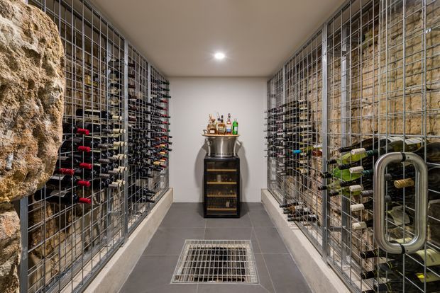 A narrow, purpose-built wine cellar lined on both sides with extensive metal wine racks and bottle storage. Rough natural stone feature walls frame the room, while dark grey floor tiles, a central floor grille, and a compact drinks cabinet with bottles create a refined cellar setting.