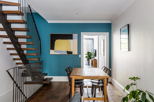 A bright, narrow dining area with a substantial timber dining table and dark metal chairs, positioned beside an open-riser staircase with timber treads and black metal balustrading. The room features a bold teal accent wall with abstract artwork, white walls and detailed cornices, dark timber flooring, and a glazed doorway leading to a brighter adjoining space. Indoor plants add a natural touch.
