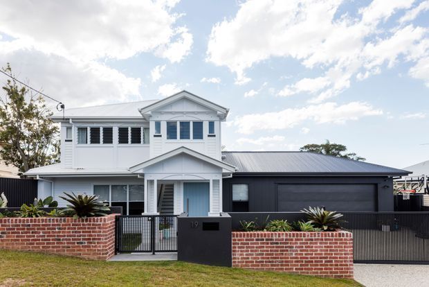 Front exterior view of a renovated or newly built suburban residence, combining a white two-storey traditional-style house with a dark single-storey garage wing. The facade features weatherboard cladding, gabled roof forms, decorative entry detailing, louvred upper windows, and a low red-brick boundary wall with landscaped planting.