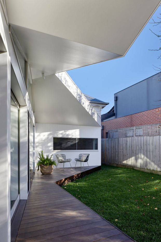 A bright, modern residential courtyard viewed from a timber deck walkway. The space features crisp white walls, a large projecting flat-roof canopy, extensive glass doors, a horizontal window, dark timber decking, lawn, and a timber-and-metal boundary fence. Two wire-frame chairs and a woven planter create a small outdoor sitting area. Neighbouring buildings and clear sky are visible beyond the courtyard.