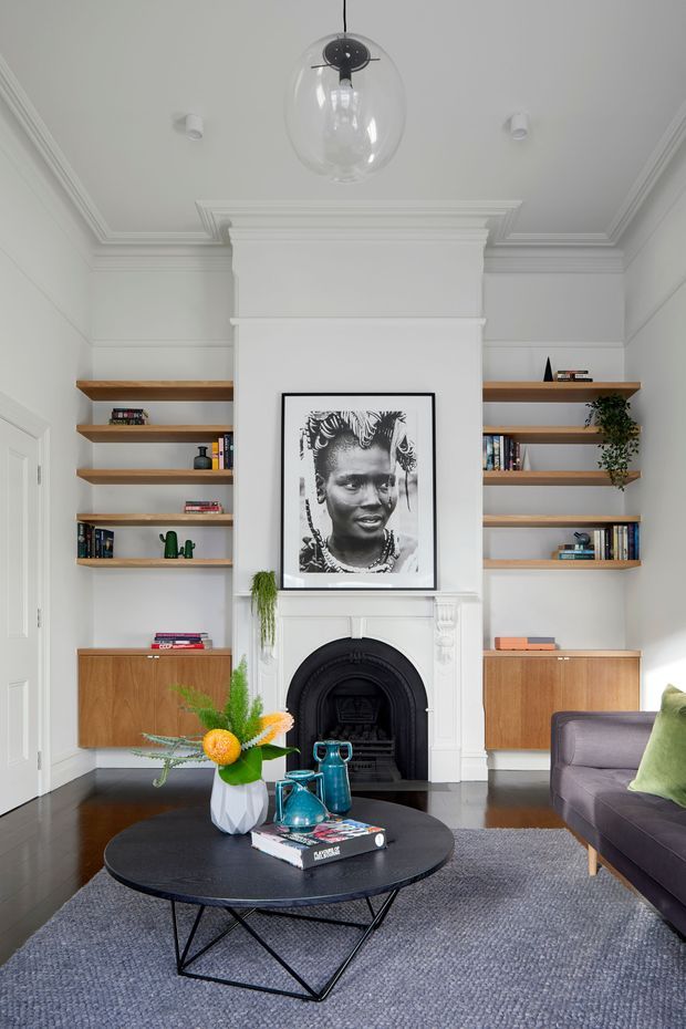 A refined living room with a central white decorative fireplace and mantel, framed monochrome portrait, and symmetrical open timber shelving with integrated wood cabinetry. The room has ornate ceiling cornices and wall mouldings, white painted walls, dark timber flooring, a grey area rug, a round black coffee table, and a dark upholstered sofa. Contemporary furniture and colourful ceramic accessories add contrast to the classic architectural detailing.