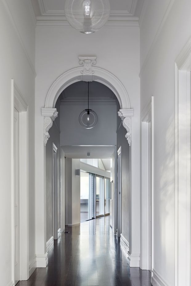 A tall, refined interior hallway with white walls, detailed traditional mouldings, ornate carved archway and pilasters, panelled doors, polished dark timber flooring, and spherical pendant lights. The corridor leads toward a brighter modern glazed area, creating a contrast between heritage detailing and contemporary openness.