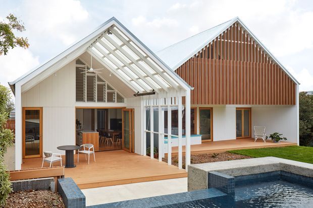 Exterior view of a contemporary residential house with two steep gabled volumes, white vertical cladding, warm timber screening and decking, extensive glazed openings, and a covered outdoor entertaining deck beside a swimming pool. The open central gable creates a bright indoor-outdoor connection, with living and dining areas visible inside.