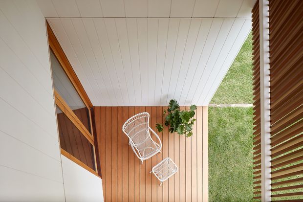 A small covered outdoor terrace or porch viewed from above, featuring warm timber decking, a white slatted ceiling, white wicker lounge chairs, potted greenery, timber-framed glazing, and a vertical timber privacy screen. The space has a simple indoor-outdoor connection and a warm natural palette.