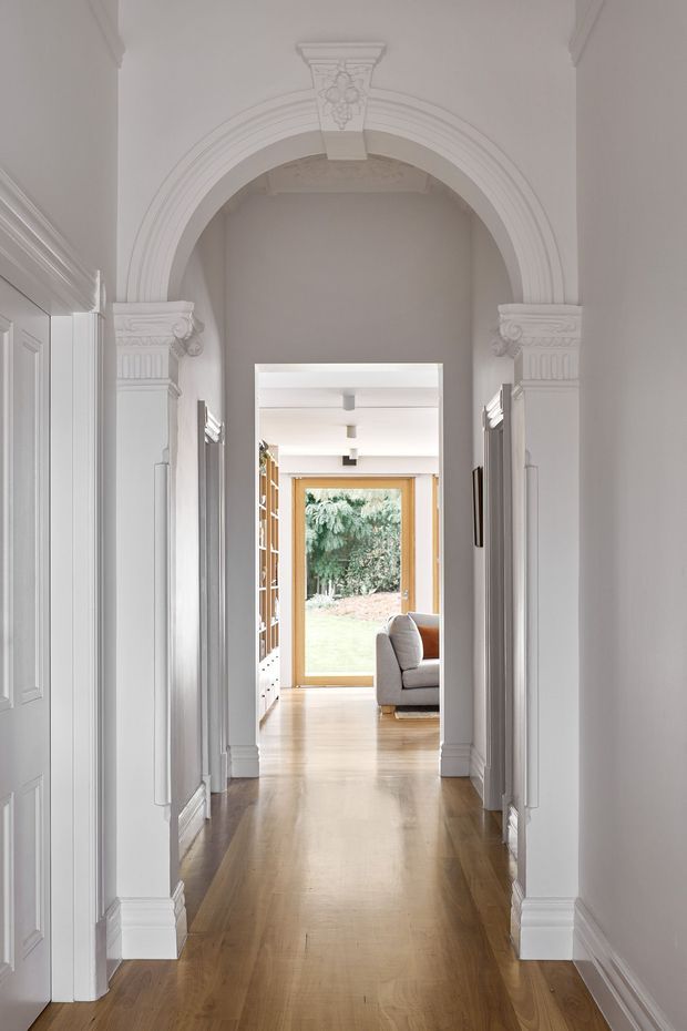 A refined residential hallway viewed through a sequence of ornate arched openings. The corridor has warm timber flooring, white painted walls, decorative classical columns and moulded architraves, panelled doors, and built-in detailing. At the far end, a large timber-framed glazed opening provides a view to greenery, with part of a contemporary living area visible beyond.