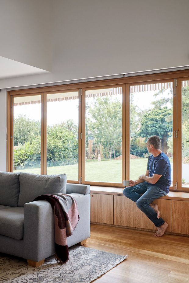 A bright, warm living room with a large grey sectional sofa, patterned area rug, and a built-in timber window seat beneath expansive timber-framed glazing. The room opens visually onto a leafy garden and lawn, with a seated person on the window bench. The space has a clean, contemporary feel, natural timber flooring and joinery, white walls, and abundant daylight.