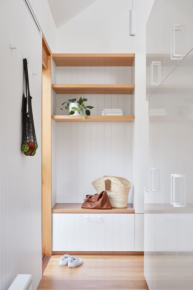 Bright, refined entryway with custom white vertical-panel joinery, open timber display shelves, a built-in bench, and concealed white cabinetry. Pale timber flooring and warm timber trim contrast with the white walls and cabinetry. A woven basket, leather bag, shoes, hanging reusable produce bag, and potted plant add understated natural accents. A tall mirrored or glossy cabinet surface is visible on the right, reflecting the entry joinery and pendant lighting.