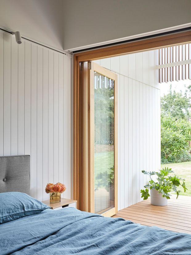 Bright, contemporary bedroom opening onto a timber deck through large glazed doors. The room features white vertical wall panelling, a grey upholstered bedhead, blue linen bedding, a small timber bedside table, and natural timber window and door framing. A potted plant and garden are visible outside, with vertical timber screening above the opening.