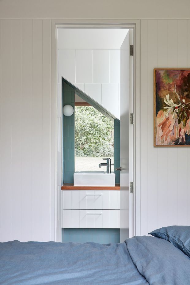 A bedroom view looking directly toward an open ensuite bathroom. The foreground shows a bed with blue bedding, while the compact bathroom beyond features a white vessel basin, chrome wall-mounted tap, white vanity cabinetry, pale blue-green wall tiles, and a timber benchtop. White vertical wall panelling surrounds the doorway, and a colourful framed artwork hangs beside it. A distinctive angled mirror or glazed opening reflects greenery outside, bringing natural light into the bathroom.