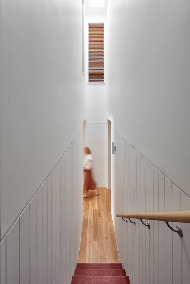 A bright, narrow stairway viewed from above, with white walls, slim vertical metal balusters, a timber handrail, and warm timber flooring on the landing below. Red carpet covers the stairs, and a tall narrow window with timber horizontal louvres brings light into the stairwell. A blurred person is visible on the landing.