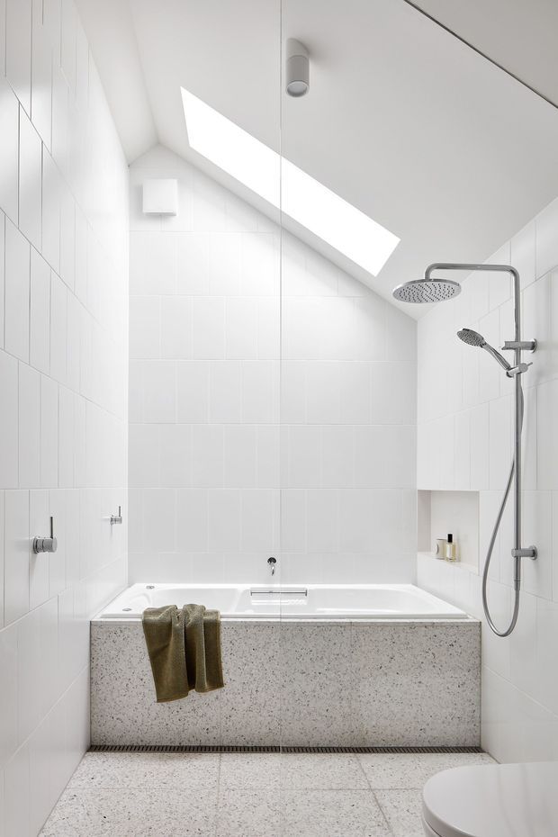 Bright, compact contemporary bathroom with a built-in white bathtub and overhead rain shower beneath a sloped ceiling. White tiled walls, a large skylight, chrome shower fittings, terrazzo-clad bath surround, and terrazzo floor create a clean, minimalist feel. A partial toilet is visible in the foreground.