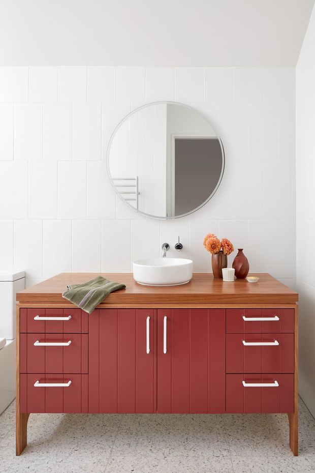 A bright, compact bathroom featuring a freestanding red vanity with white handles and a warm timber benchtop. A white vessel basin sits beneath a large round mirror, with white tiled walls, terrazzo-style flooring, and minimal decorative styling including ceramic vessels and orange flowers.