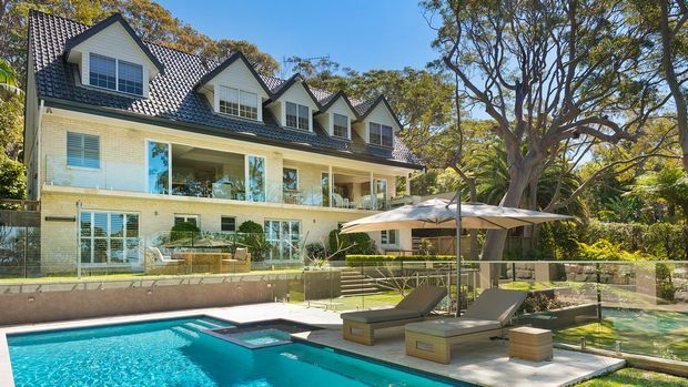A large multi-storey suburban residence with a steep tiled gabled roof, multiple dormer windows, pale brick walls, extensive glazed verandah-style balconies, and white-framed windows. The landscaped backyard includes a swimming pool, spa, glass balustrade, outdoor lounge chairs, and a large shade umbrella beneath mature trees.